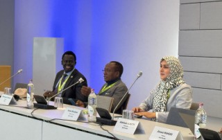 Three people sit in front of microphones at a board meeting.