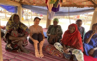 A group of women sit cross-legged on woven mats under a patterned fabric shelter, wearing colorful clothing and headscarves, gathered closely together in a shaded outdoor setting.